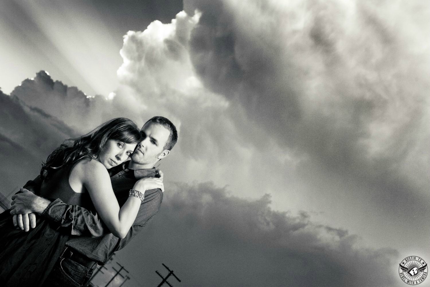 male and female couple wearing trendy clothes  embrace and she has curly dark hair in dark dress and he has short dark hair with a dark button up shirt in front of dramatic ominous clouds with rays of sunshine coming from behind the cloud  